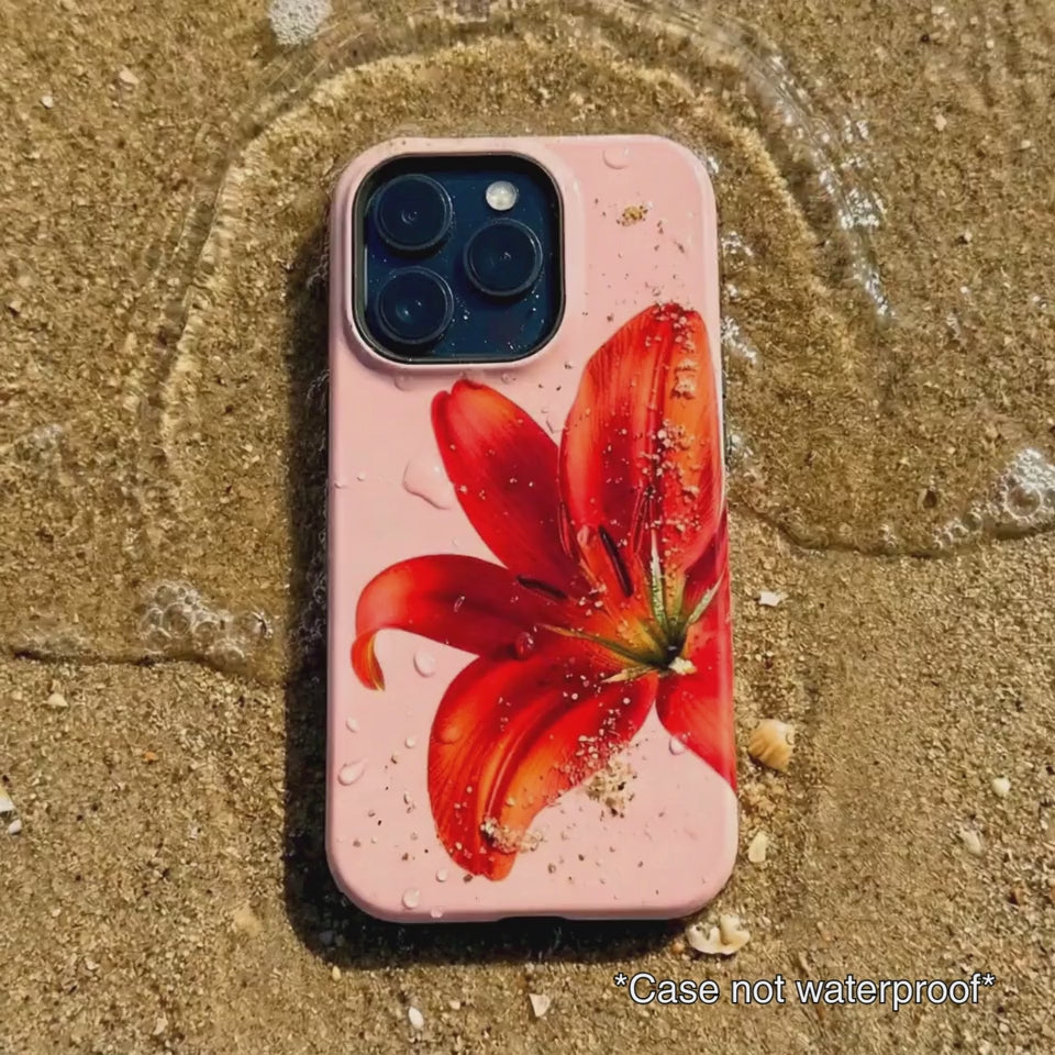 pink iPhone case with red floral design sitting on beach sand as waves gentle wash over it.