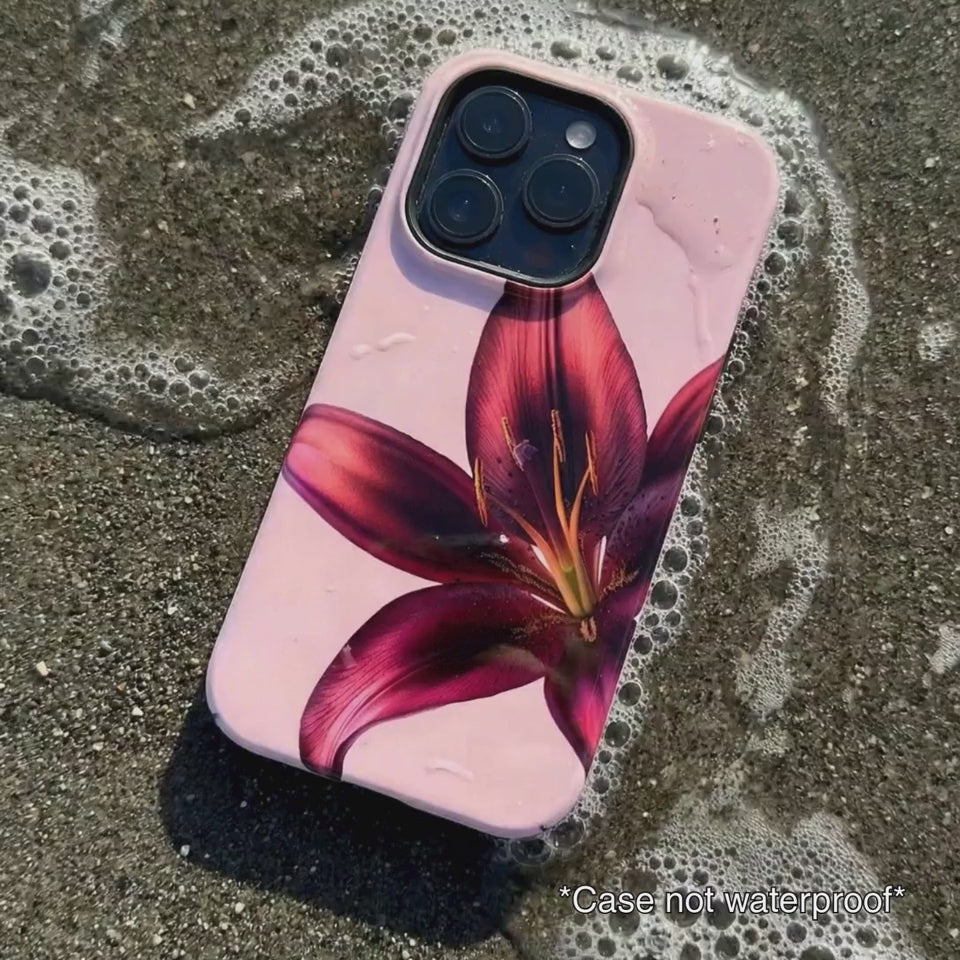 pink iPhone case with purple floral design sitting on beach sand as waves gentle wash over it.