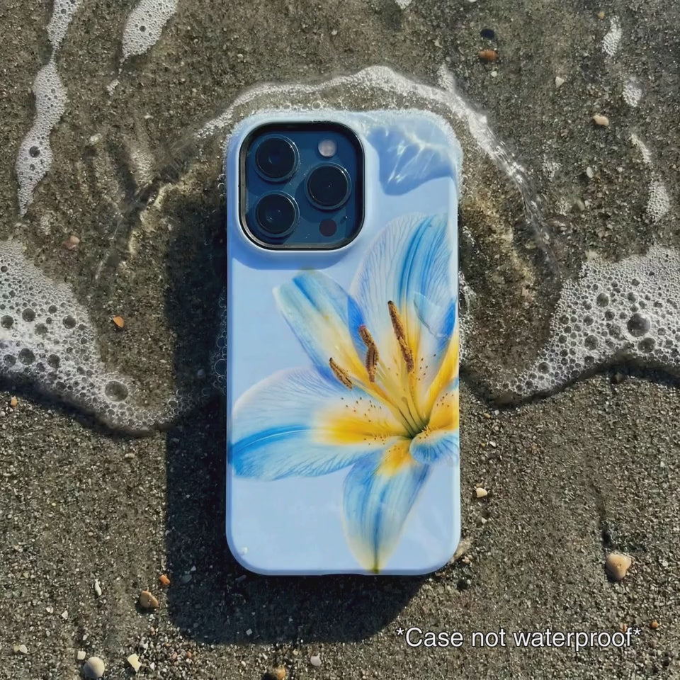 light blue iPhone case with floral design sitting on beach sand as waves gentle wash over it.