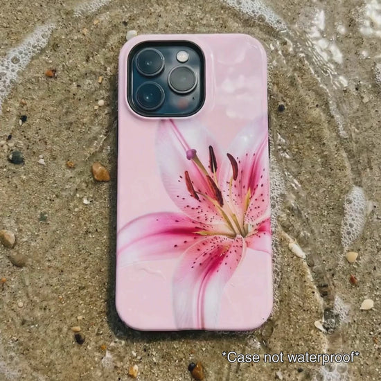 pink iPhone case with floral design sitting on beach sand as waves gentle wash over it.