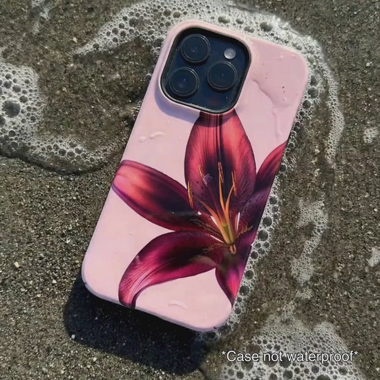 pink iPhone case with purple floral design sitting on beach sand as waves gentle wash over it.